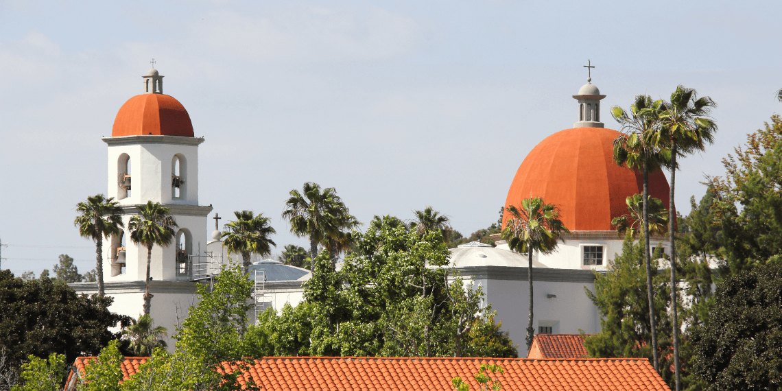 Scenic view of San Juan Capistrano's iconic church with distinctive red roofs, set against a backdrop of lush green trees, blending architectural beauty with natural serenity.