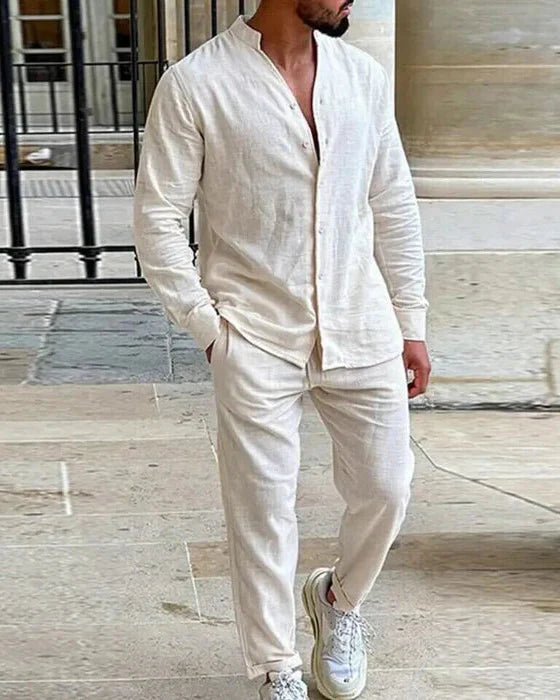 A man wearing an off-white linen set consisting of a long-sleeved shirt with a stand-up collar and long trousers stands in a stone-tiled courtyard.