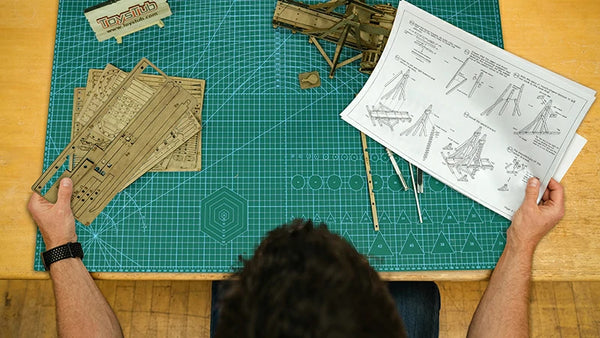 a man holding wooden puzzle parts board in his left hand and instrction in his right hand, sitting on a table with a cutting mat on it.