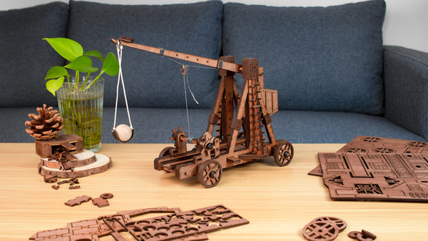 a 4 wheels counteweight trebuchet wooden model lying on a coffee-table with it's parts boards and a sofa in background