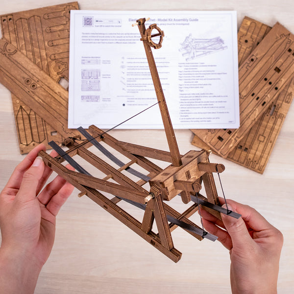 two hands holding the Horizontal Trebuchet model with instruction and wooden parts in background