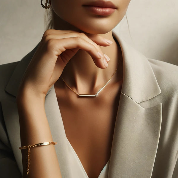 A stylish photograph focusing on a woman from the neck down, showcasing her minimalist bar gold necklace and a minimalist chain gold bracelet as she poses elegantly with her hand under her chin. The soft, neutral setting highlights the delicate and simple elegance of the jewelry