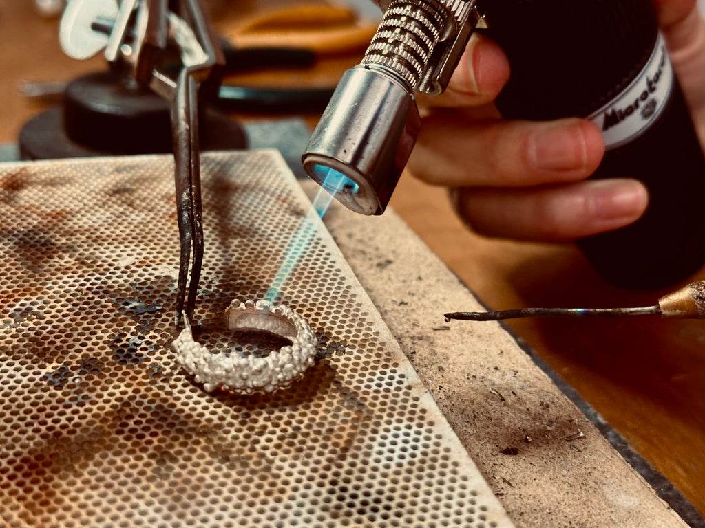 Artisan at What If You Stayed using a torch to solder a textured sterling silver ring in their Montreal workshop.