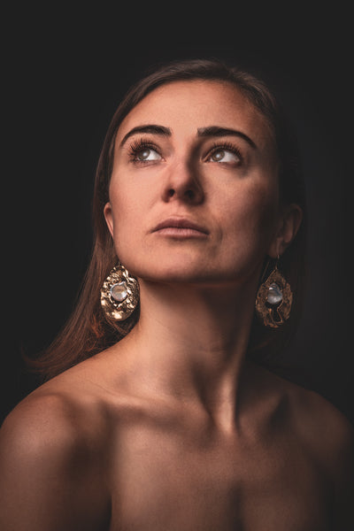 Woman with a thoughtful gaze wearing 'What if you stayed' ornate gold statement earrings, featuring iridescent gemstones, against a dark backdrop