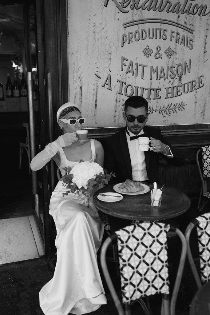 A cool bride drinks coffee with her groom in Paris street cafe wearing a slim fitting wedding gown and a birdcage veil headband