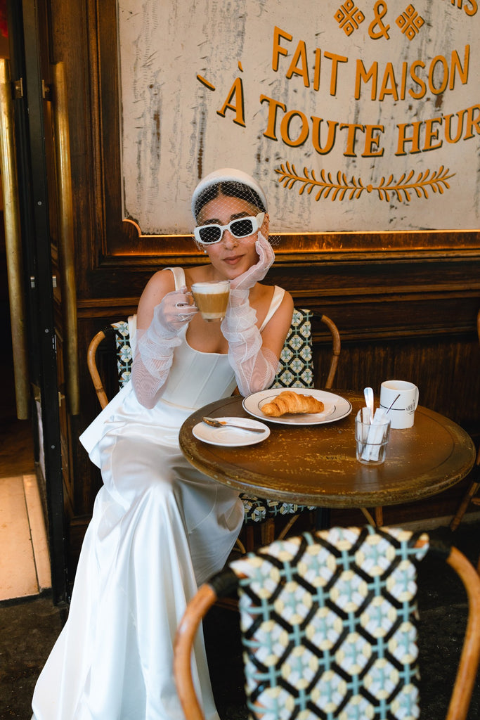 A cool bride wears a birdcage veil headband with white sunglasses in a Paris cafe
