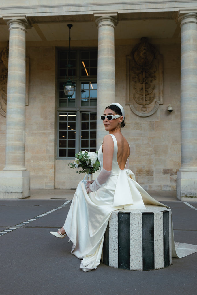 A cool bride sits on a black and white striped circular block in Paris in a modern white gown and a padded headband with a birdcage veil