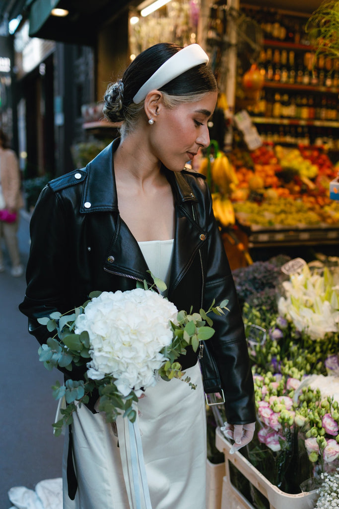 A cool bride wears a black leather jacket over her slim fitting wedding gown, carrying a bouquet and wearing a birdcage veil headband