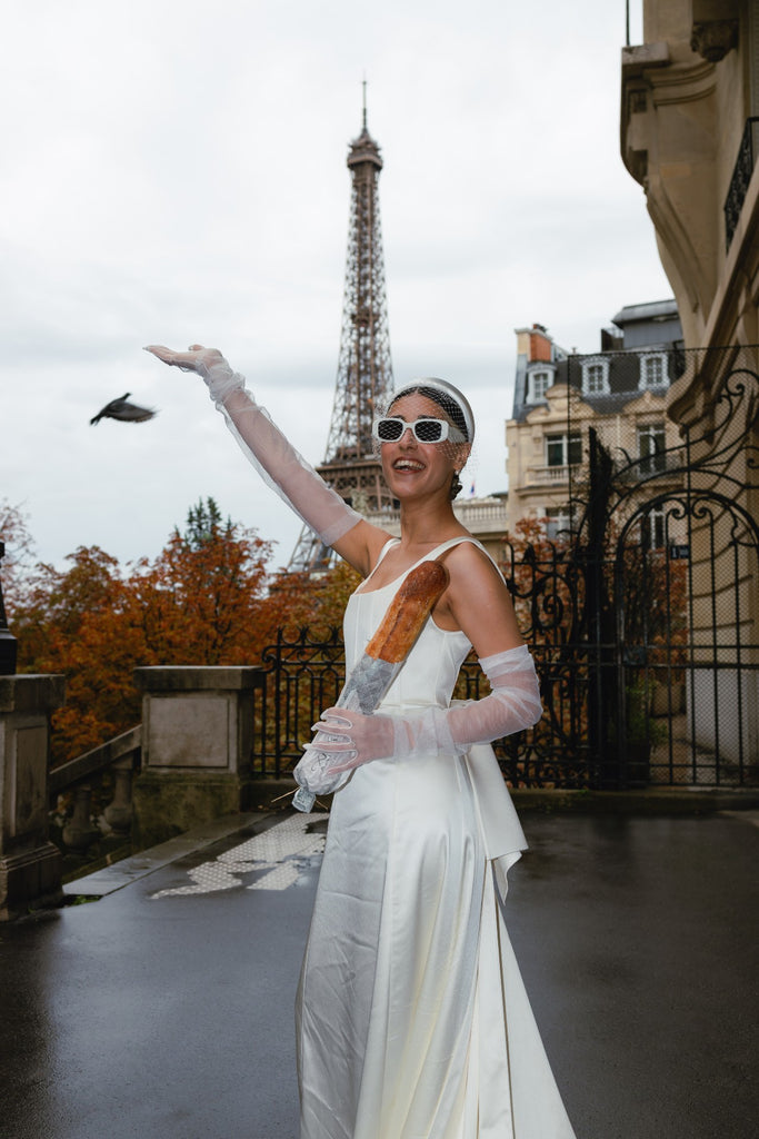 A bride poses in front of the Eiffel Tower in a birdcage veil headband and white framed sunglasses with her arm raised in the air and carrying a baguette