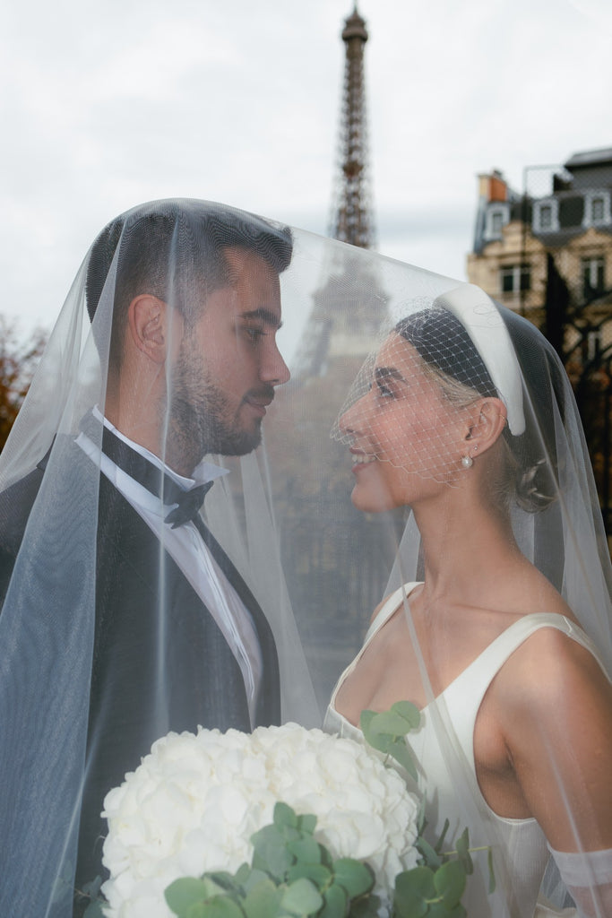 A cool bride wears a birdcage veil headband in front of the Eiffel Tower in Paris and poses with her groom under a gauzy long wedding veil