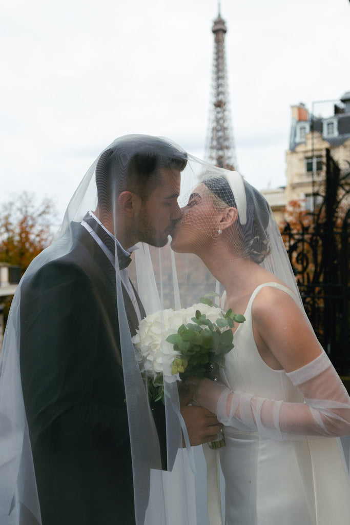A cool bride wears a birdcage veil headband in front of the Eiffel Tower in Paris and kisses her groom under a gauzy long wedding veil
