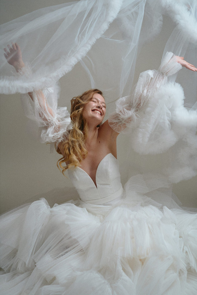 Bride sits in a sea of tulle and throws a veil up in the ear with a look of joy - Debbie Carlisle 2024 collection shoot image