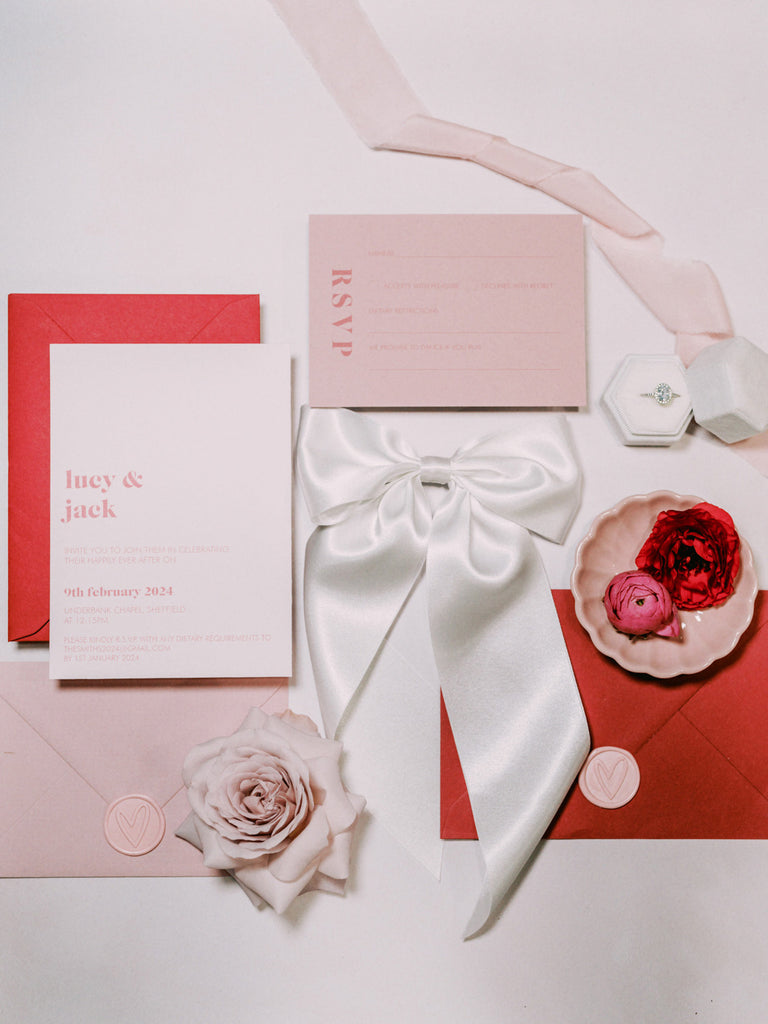 A white satin hair bow and pink and red wedding stationery for a modern romantic bride and groom
