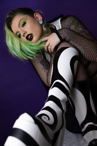 Alt model Naomi wearing the nightshade black liquid lipstick and Killin it Cemetery gree eyeshadow palette and wearing black and white swirl boots from Current Mood and Dolls Kill against a purple backdrop