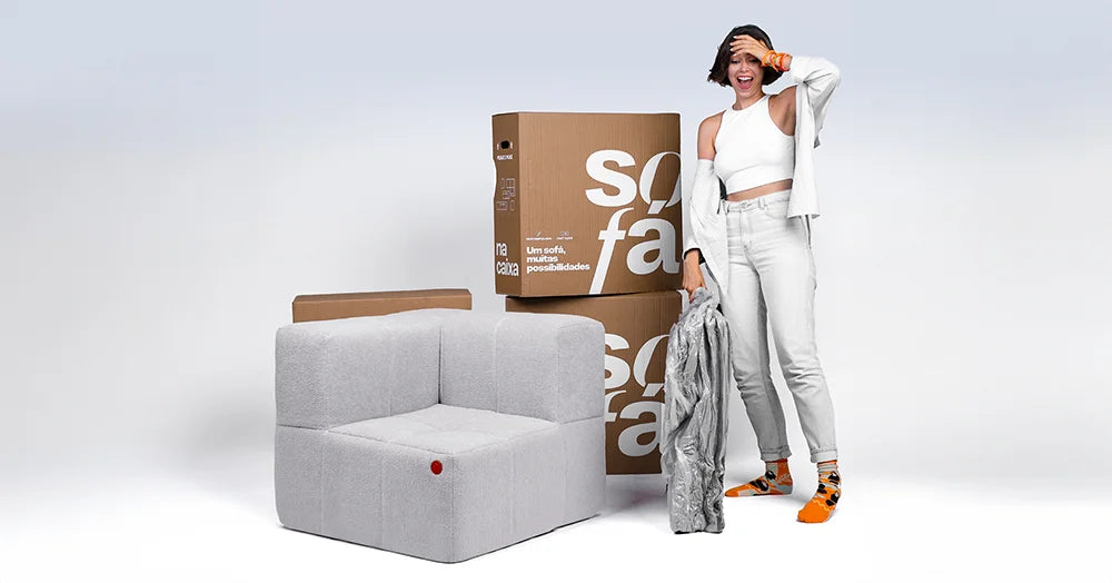 Mulher sorridente ao lado de um sofá modular cinza com um botão vermelho, com caixas de papelão marcadas com 'sofa' ao fundo, sugerindo um produto de mobiliário que pode ser facilmente montado e personalizado.