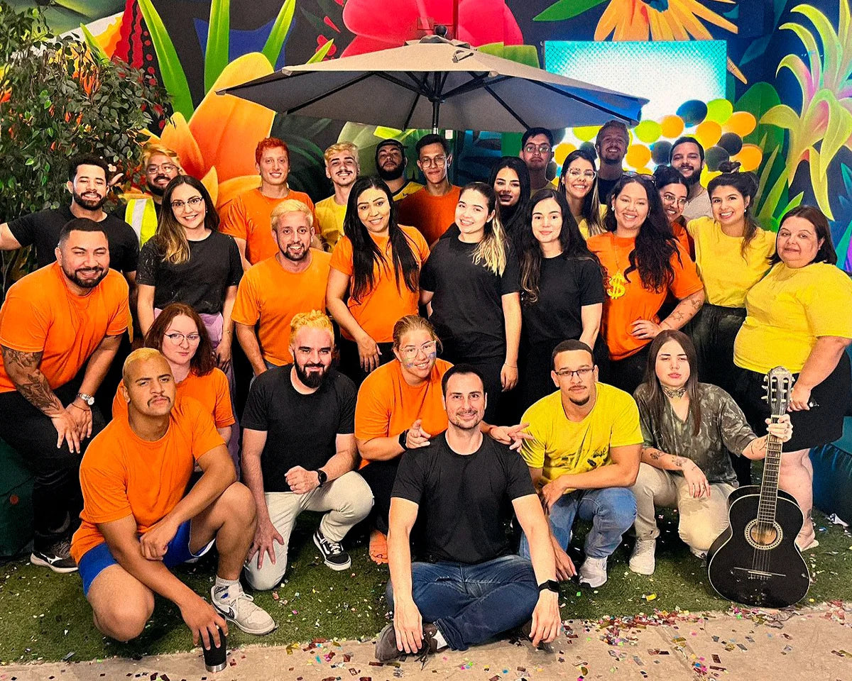 Grupo de trinta pessoas sorridentes posando para uma foto em um ambiente com uma pintura indígena nas paredes em cores vivas como amarelo, vermelho e azul. Doze mulheres e dezoito homens estão vestidos com roupas casuais, muitas em tons de laranja, preto e amarelo. Eles estão em pé em uma semicircunferência. A atmosfera é descontraída e alegre, com as pessoas sorrindo para a foto.