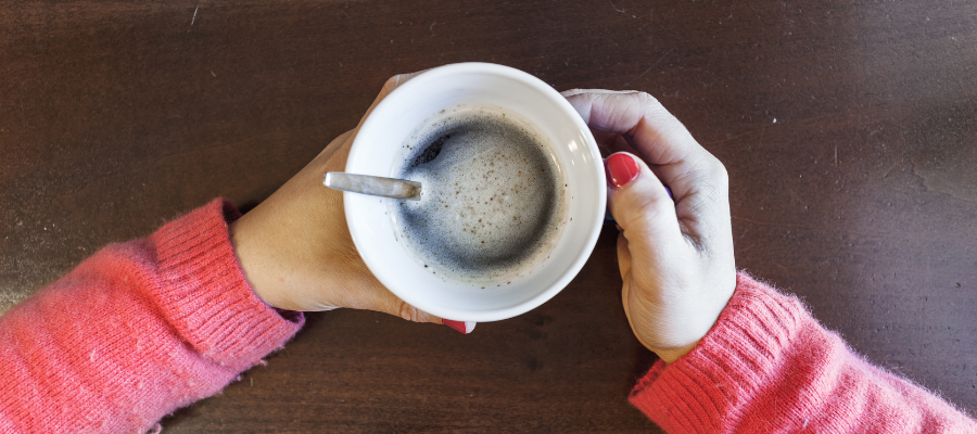 woman wearing pink jumper holding a black coffee in a white mug on a brown table