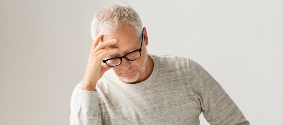 white man with grey hair and black glasses looking down and holding his head in stress