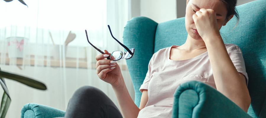 Lady sat on blue chair with pink white top, covering eyes in tiredness and fatigue, holding her glasses