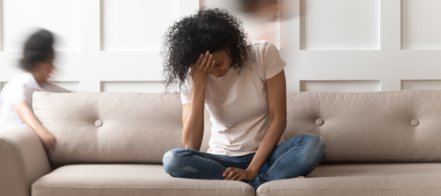 woman with black afro hair covering her face whilst sat on a cream sofa with children running around