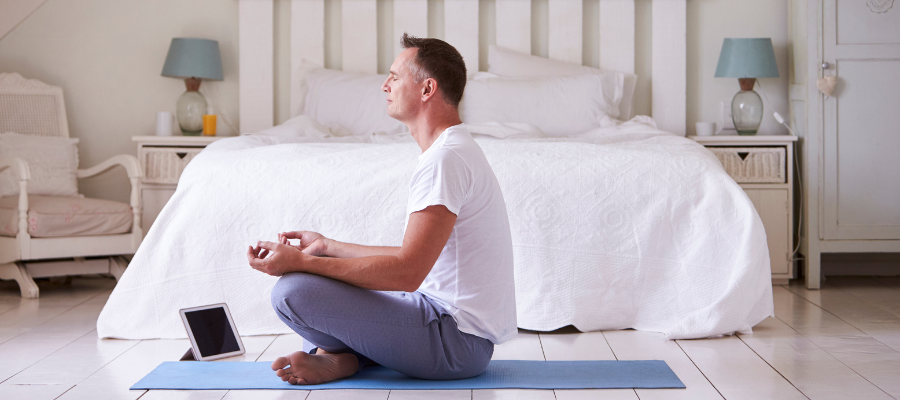 man with grey hair in bedroom meditating to an app on his iPad on a yoga matt