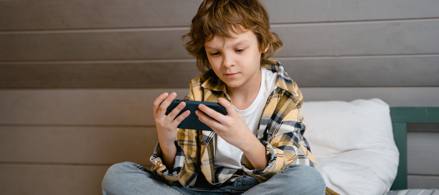 young boy playing on his phone on a bed wearing a yellow and grey shirt