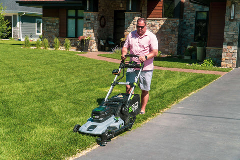 A middle-aged man is mowing and edging the front yard with a push lawn mower and a Trimyxs attached