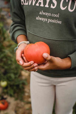 Girl wearing green sweater and beige pants holding a large, red tomato