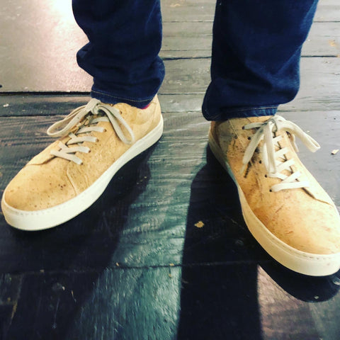 A close up of a man wearing the cork sunslider kicks sneakers on a black wood floor.