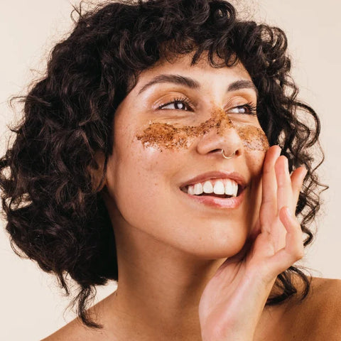 A woman applying brown exfoliating scrub to her face, showcasing a skincare routine for healthy, glowing skin.