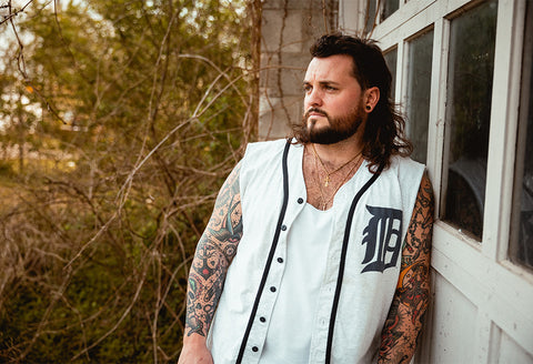 Country-rock artist Jason Cross in a cutoff baseball jersey leaning on an abandoned building.