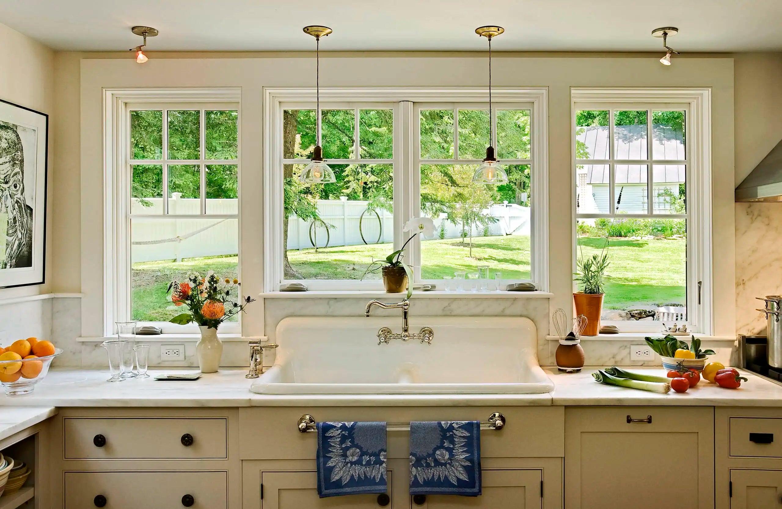 A bright farmhouse-style kitchen featuring a large porcelain utility sink under three windows with garden views. | ACE DECOR