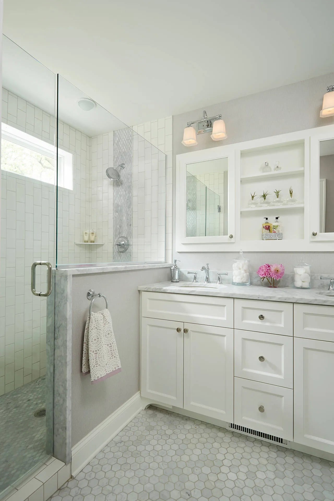 White wood bathroom cabinet with marble countertop, subway-tile shower, hexagonal floor tiles, pink floral accents, and round metal knobs storage.