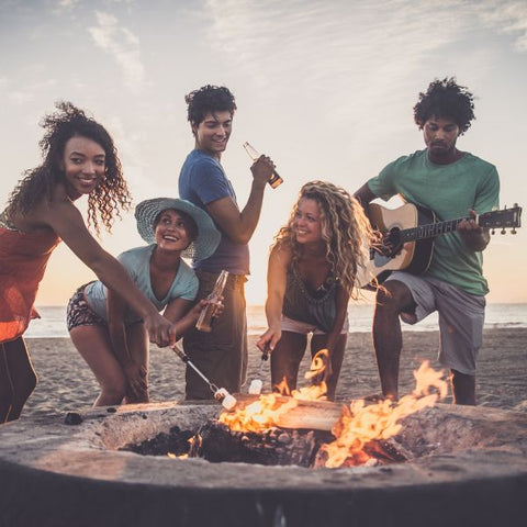 Friends gather around a beach bonfire, roasting marshmallows and playing guitar
