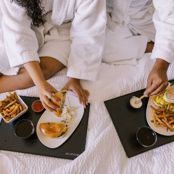 A couple in white robes enjoys a hotel luxury of burgers and fries served on black trays in bed symbolizing indulgent comfort and relaxation.