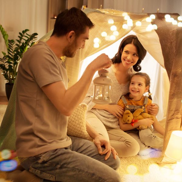 A family creating a magical indoor camping scene with a little girl holding a stuffed animal inside a blanket fort adorned with fairy lights.