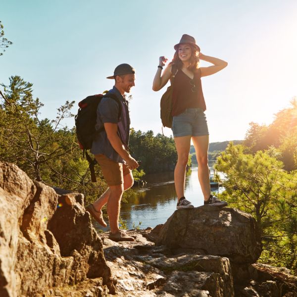 A smiling couple equipped with backpacks stands on a rocky ledge surrounded by verdant forest overlooking a calm river on a sunny day reflecting a sense of adventure and discovery.