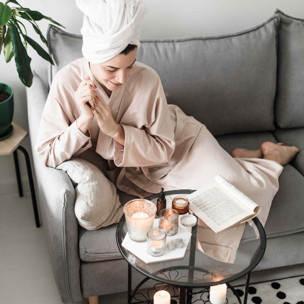 A woman in a cozy robe and towel-wrapped hair sits on a gray couch with a book surrounded by candles and skincare products for a tranquil self-care routine.