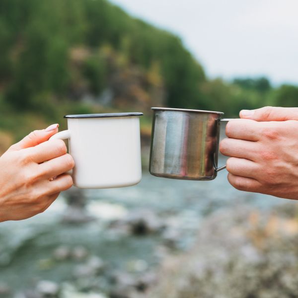 Two hands clinking a white enamel mug against a stainless steel cup in the foreground with a blurred river and greenery in the background.