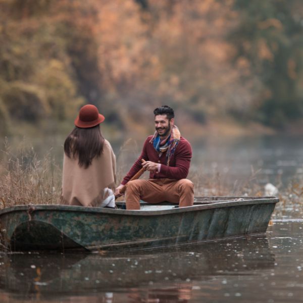 A casual yet romantic outdoor scene depicting a man in a maroon sweater and brown pants sharing a laugh with a woman in a brown hat and beige coat.