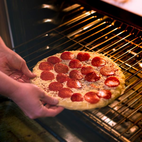 A freshly made pepperoni pizza with vibrant red slices of pepperoni and melted cheese being placed into an oven by a person's hands showcasing homemade cooking.