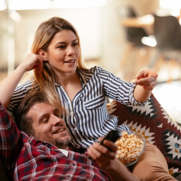 A couple enjoys a cozy moment on a couch with the man holding a remote and the woman playfully pointing while they share a bowl of popcorn.