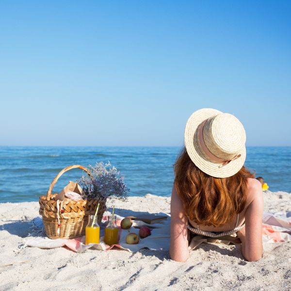 Woman lying on a beach towel next to a picnic basket and fruits on the sandy beach