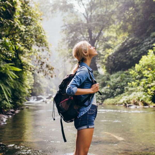 A young woman with a backpack stands in a shallow river surrounded by a dense tropical forest looking up with a sense of wonder and adventure.