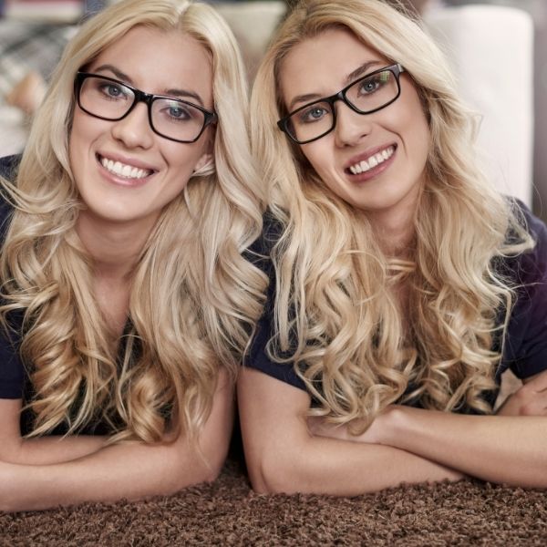 Smiling blonde twin sisters wearing glasses and lying on the carpet.