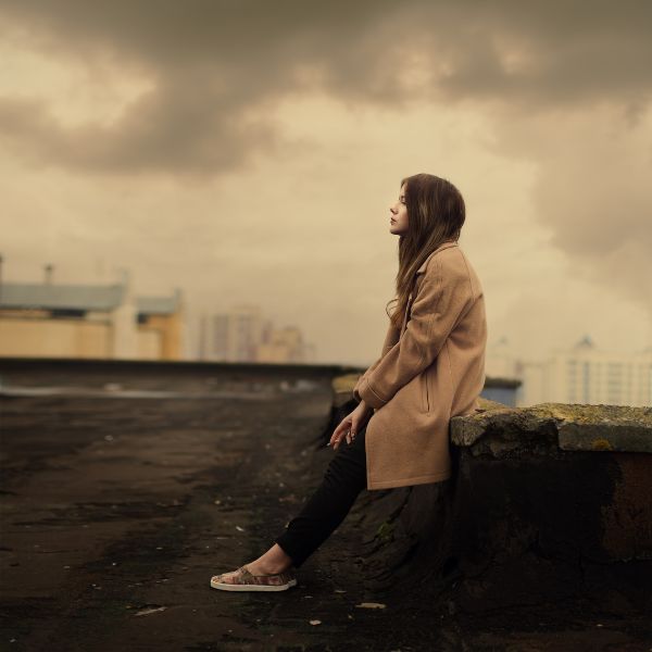 A young woman sitting on a rooftop ledge, looking out over the city under a dramatic sky
