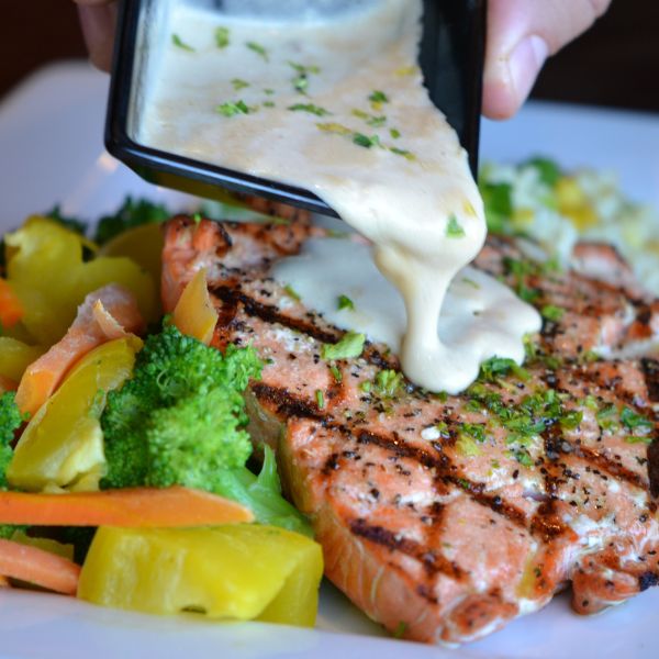 A hand pouring sauce over a grilled salmon fillet surrounded by vegetables, showcasing a delicious and healthy meal being plated.