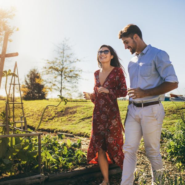 A cheerful couple enjoying a sunny day in a garden with the woman in a flowing red patterned dress and the man in a light blue shirt and white pants strolling and laughing together among the greenery.