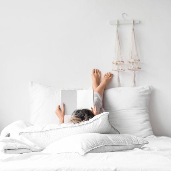 View from the foot of a bed showing a person lying down with a book open upside down evoking a relaxed and carefree atmosphere in a minimalist bedroom