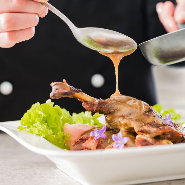 A succulent roasted duck leg being basted with a spoonful of glaze beside a bed of lettuce and tomatoes highlighting gourmet cooking and presentation.